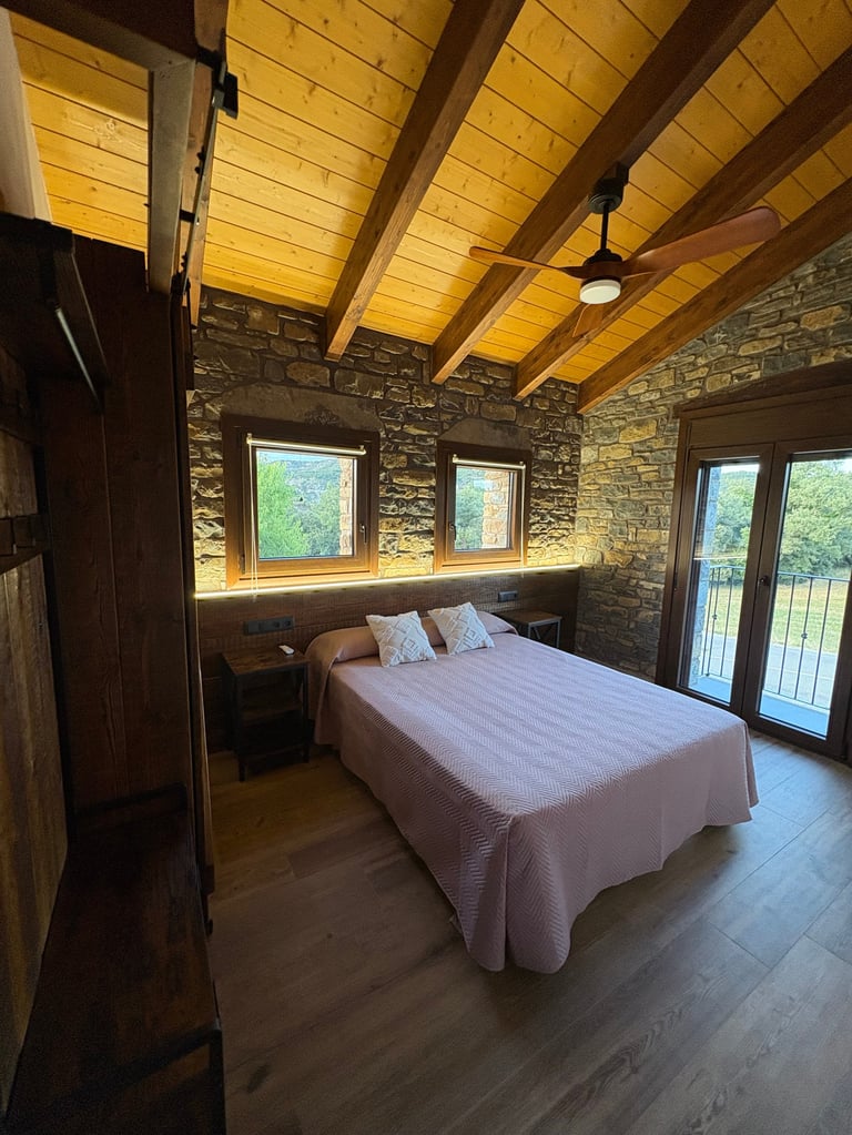 Modern bedroom with wooden beamed ceiling, exposed stone walls, queen bed with white bedding, and glass doors leading to a balcony overlooking greenery