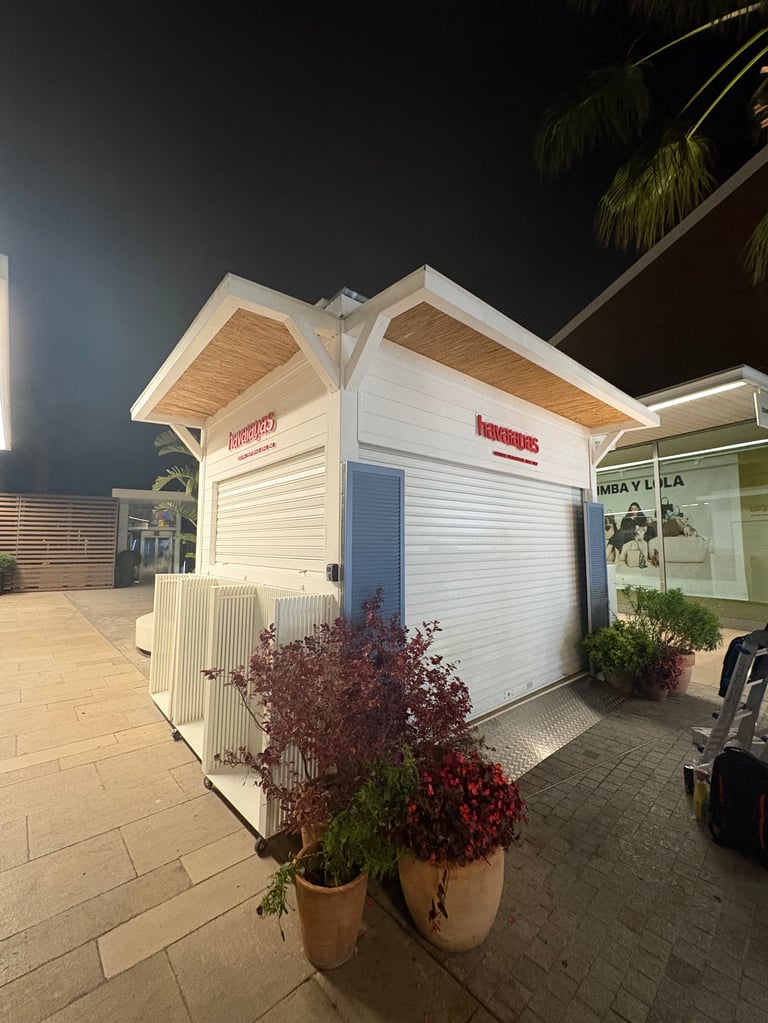 White beach hut with peaked roof at night, flanked by potted burgundy plants and palm trees nearby