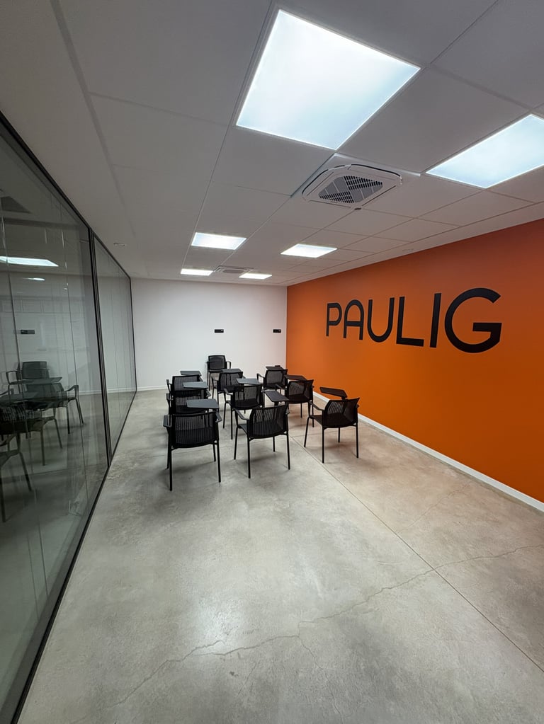 Modern office meeting room with orange accent wall displaying PAULIG logo, black chairs, glass partition, and skylight ceiling panels