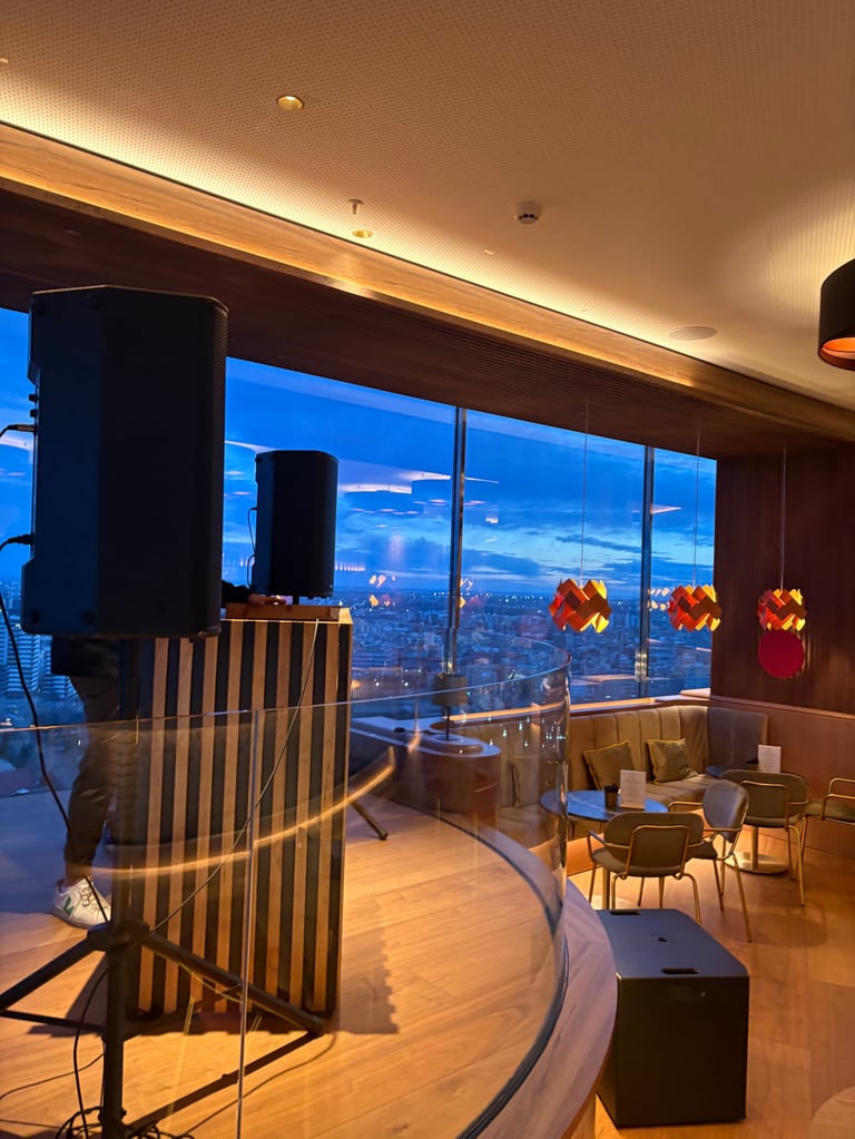 Modern high-rise lounge with floor-to-ceiling windows overlooking a city skyline at dusk, featuring wood flooring, contemporary seating, and ambient lighting